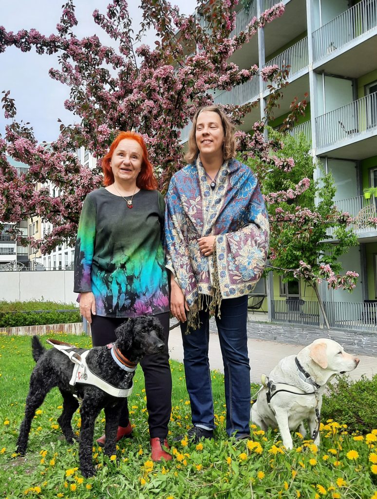 Riikka ja Iiris seisovat koirien kanssa puun juurella.